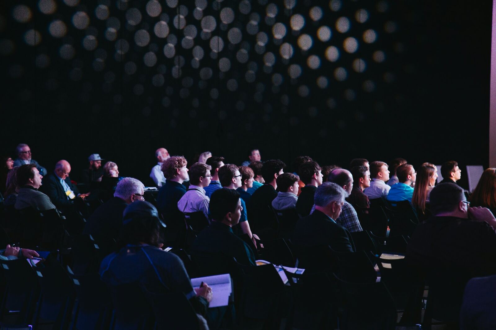 An audience seated in a darkened auditorium, stage lights out of focus behind them