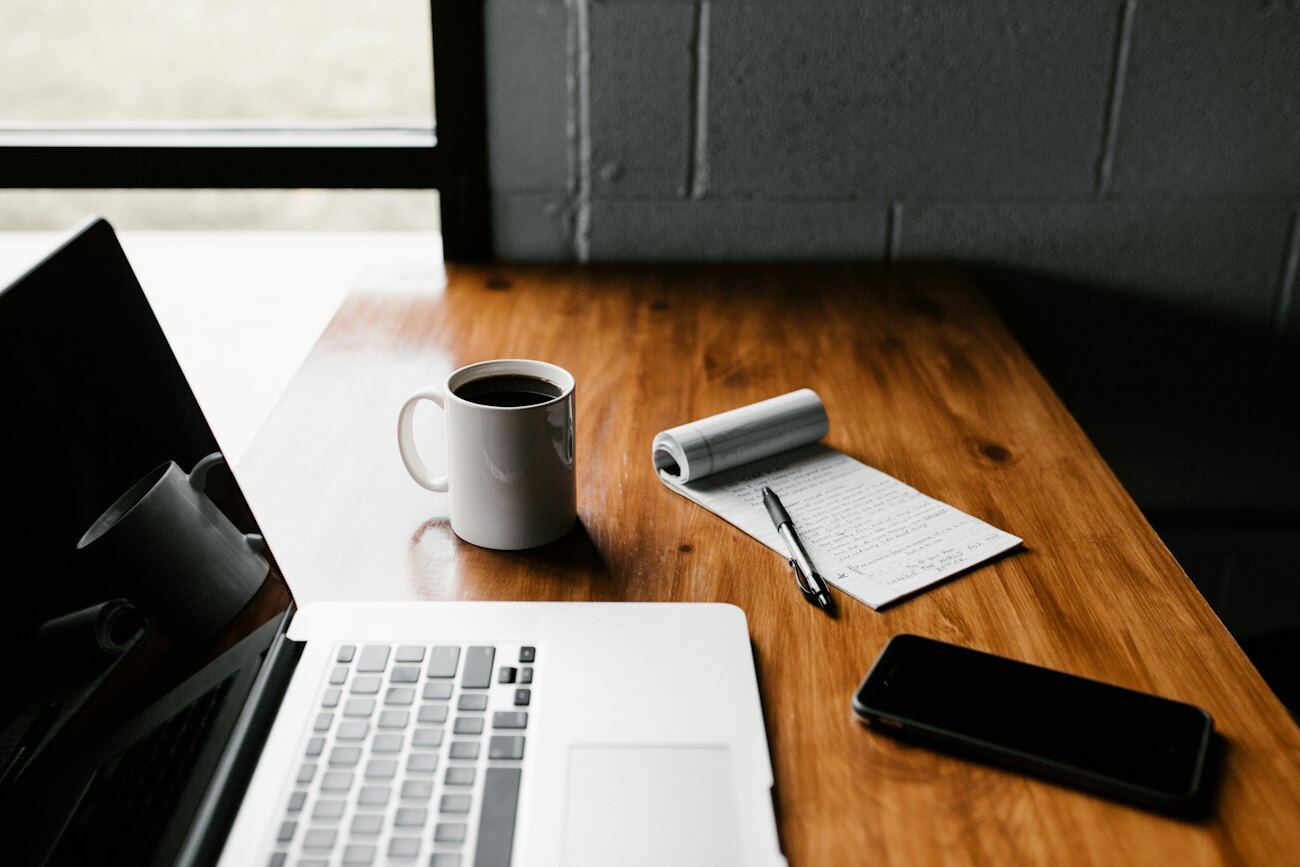 A desk with an open notebook, a pen, a laptop and a cup of coffee